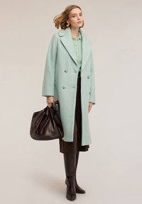 Abrigo cruzado de doble botonadura en verde claro texturizado, con solapas grandes y botones plateados, combinado con un bolso marrón, una falda marrón y botas marrones hasta la rodilla.