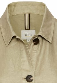 Beige skjorte med en struktureret krave, der har en brun knap, og et mærke, der viser "camel active." Tekstureret stof med en subtil glans.