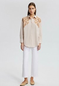 Cream-colored blouse with a textured fabric featuring intricate brown embroidery on the shoulders. Paired with white cropped jeans and beige flats.