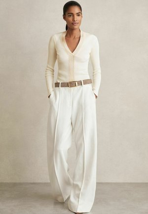 Top a maniche lunghe a coste color crema con scollatura a V e dettaglio in catena dorata, abbinato a pantaloni bianchi a gamba larga e cintura in pelle marrone. Tessuti lisci.