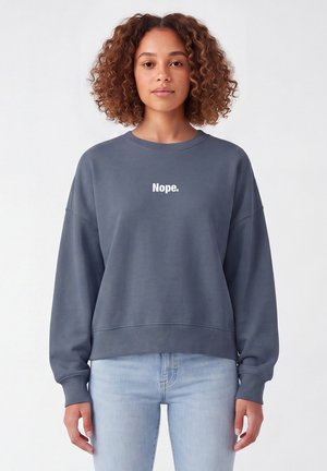 Junge Frau mit lockigem Haar, die einen blauen Sweatshirt mit dem Wort "Nope." und hellblaue Jeans trägt, steht vor einem weißen Hintergrund.