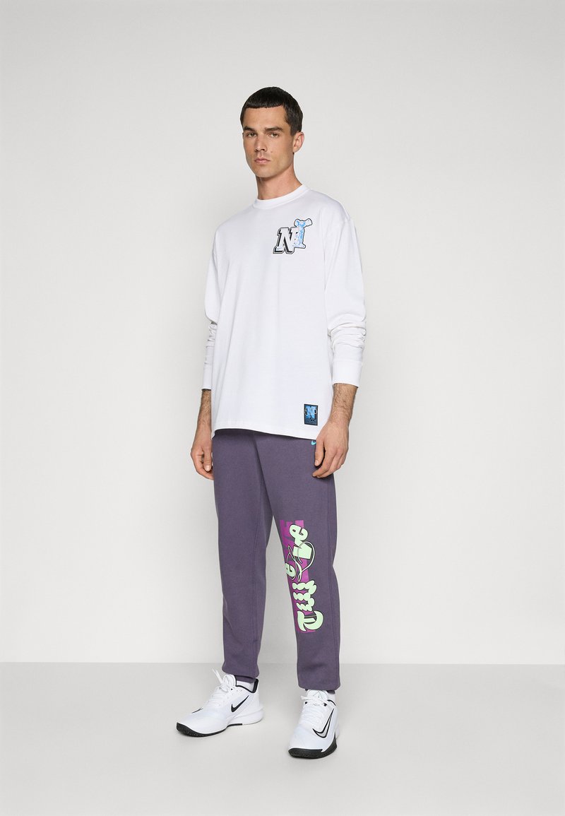 T-shirt blanc à manches longues avec un graphisme bleu ; pantalon de survêtement gris avec un texte coloré ; chaussures de sport blanches ; arrière-plan neutre.