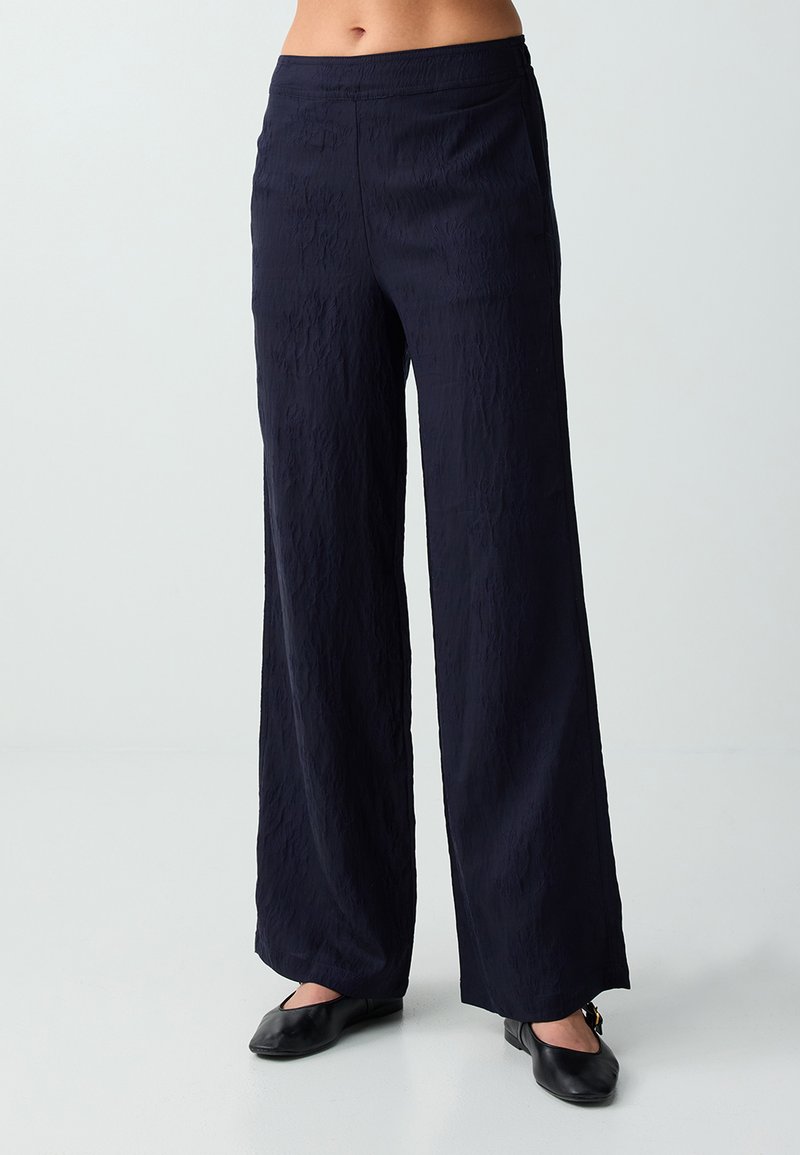 Femme portant un pantalon noir taille haute à jambes larges, associé à des chaussures plates noires, debout devant un fond clair uni.