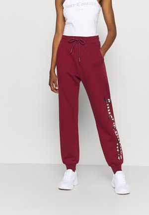 Vrouw die bordeauxkleurige joggers draagt met zwart-witte "Juicy Couture" tekst op linkerbeen en witte sneakers, staand tegen een effen achtergrond.