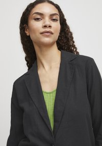 Schwarzer Leinen-Blazer mit Reverskragen, getragen über einem strukturierten grünen Oberteil. Das Model hat lockiges Haar und einen neutralen Hintergrund.