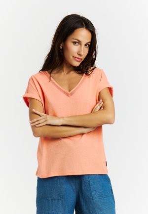 Frau mit langem dunklem Haar, die ein korallenrotes V-Ausschnitt-Kurzarmshirt und blaue Hose trägt, mit verschränkten Armen vor weißem Hintergrund steht.