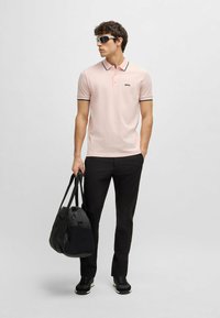 Polo shirt rosa chiaro con colletto e accenti neri sulle maniche, pantaloni neri, sneakers nere e una borsa a tracolla nera. Modello che indossa occhiali da sole.