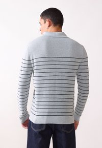 Grauer gestreifter Pullover mit strukturiertem Strick, der horizontale schwarze Streifen und einen klassischen Kragen aufweist. Getragen mit dunklen Jeans.
