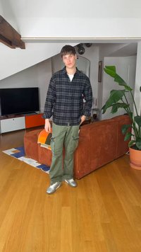 Νεαρός άνδρας με πράσινο παντελόνι cargo και σκούρο πουκάμισο καρό στέκεται σε ένα μοντέρνο σαλόνι με πορτοκαλί καναπέ, τηλεόραση και μεγάλο γλαστράκι.