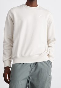 Light beige sweatshirt with a round neck and ribbed cuffs. Features a small white Nike logo on the chest. Pairing with tailored green cargo pants.