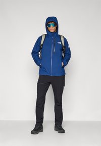 Μπλε αδιάβροχο μπουφάν με κουκούλα, μπροστινό φερμουάρ, μαύρα παντελόνια και ανθεκτικά μαύρα μποτάκια πεζοπορίας. Εξοπλισμένο με γκρι σακίδιο και γυαλιά ηλίου.