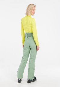 Langsleeve, felgele top gecombineerd met hooggetailleerde, lichtgroene skibroeken. De broeken hebben een geventileerde onderzijde en een slank ontwerp. Zwarte skibotten maken de outfit compleet.