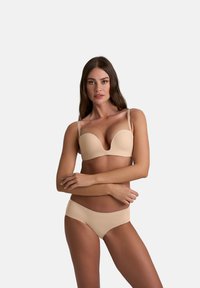 Donna con lunghi capelli castani, che indossa un reggiseno imbottito beige e una mutanda coordinata, in piedi con le braccia incrociate su uno sfondo semplice.
