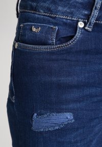 Jeans de mezclilla azul oscuro con un parche desgastado en el muslo, un pequeño bolsillo frontal y detalles de metal plateado.