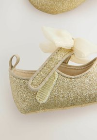 Goldene Glitzer-Balletschuhe mit einer beigen Rips-Schleife, elastischem Riemen und strukturiertem Sohlen. Der Schuh hat ein glattes Innenfutter und einen dezenten Schimmer.