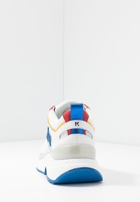 Vit sneaker med blå sula, röda och blå accenter, samt "Karl Lagerfeld"-text på hälen, visad bakifrån på en vit yta.