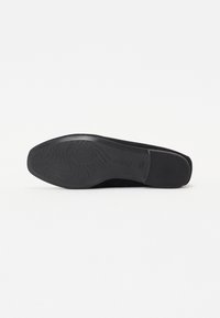 Zwarte suède platte schoen met een rubberen zool en een golvend profiel. De schoen heeft een ronde neus en een gladde afwerking.
