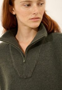 Mörkgrön tröja med dragkedja i kragen; har ribbad textur och en delad V-halsdesign. Mjukt material med en jämn finish.