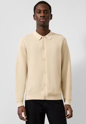 Man with dark skin wearing a beige buttoned knit cardigan and black pants, standing against a plain light background.