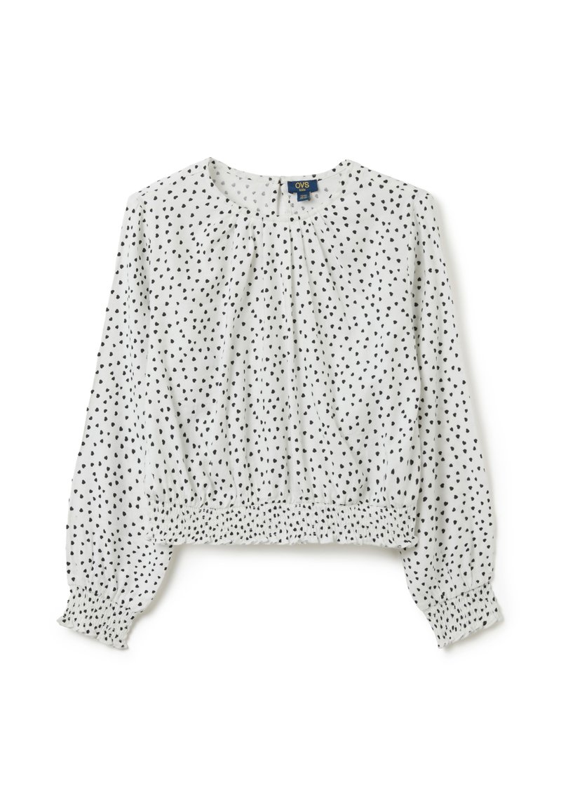 Witte kinderblouse met lange mouwen, voorzien van een zwart onregelmatig stippenpatroon, elastische manchetten en een gathered elastische zoom.