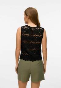 Top senza maniche in pizzo nero con motivo floreale, orlo scallop e dettaglio a chiave sul retro. Indossato sopra pantaloni corti verde oliva.