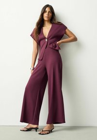 Vinrød jumpsuit med dyb V-hals, vide ben og en rynket talje. Har en dekorativ knap foran og tekstureret stof.