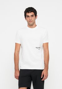 Mann med kort mørkt hår iført en hvit t-skjorte med liten svart tekst på brystet og svarte shorts, stående mot en enkel hvit bakgrunn.