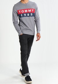 Sudadera gris con una franja horizontal roja y azul marino que presenta "TOMMY" y "1985" impresos en blanco, combinada con jeans oscuros y zapatillas negras.