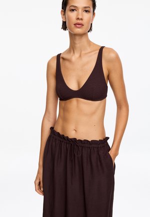 Jeune femme portant un bralette texturé marron foncé et un pantalon ample taille haute avec une ceinture froncée, sur fond blanc.