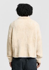 Giacca in fleece color crema con una finitura morbida e testurizzata, dotata di collo a scialle e spalle scese, indossata con pantaloni in denim scuro.