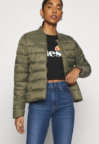 Casaco puffers acolchoado verde-azeitona com design de bomber, apresentando um fecho de correr frontal e gola canelada. Usado sobre uma t-shirt preta curta e jeans azuis.