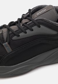 Μαύρο αθλητικό παπούτσι με υφασμάτινη υφή, λείες λεπτομέρειες και χοντρή γκρι καουτσούκ σόλα. Διαθέτει σύστημα κορδονιών και διακριτική πλευρική επωνυμία.