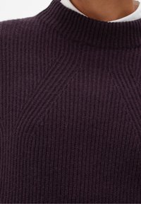 Gros plan sur un pull en tricot côtelé violet foncé avec un col rond haut, porté par-dessus une chemise blanche à col.