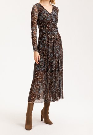 Femme portant une robe longue à manches longues et col en V, longueur cheville, avec des motifs cachemire complexes en brun, bleu et noir, associée à des bottes à talons marron.