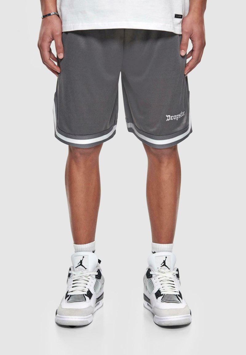 Personne portant un short de basket en mesh gris avec des rayures blanches et des baskets Air Jordan blanches et noires, debout devant un fond uni.
