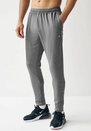 Man wearing gray athletic joggers, black and white sneakers, standing with one hand in pocket against plain white background.