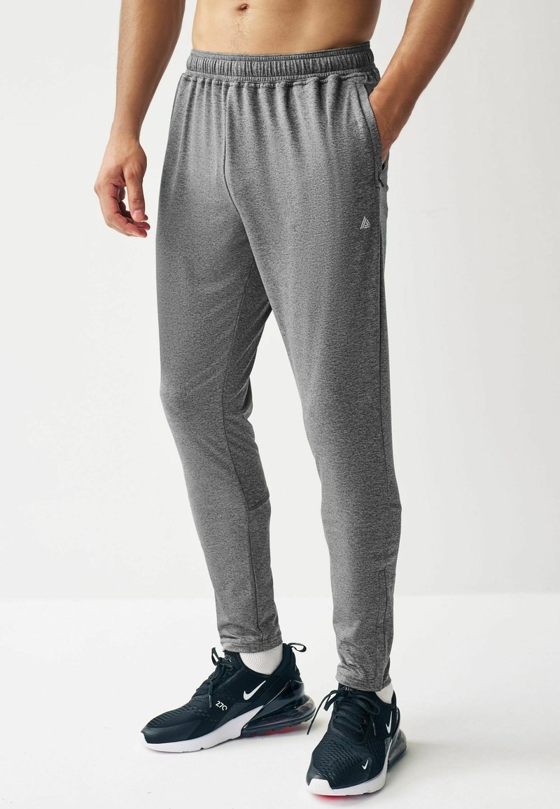 Homme portant un pantalon de jogging gris, des baskets noires et blanches, debout avec une main dans la poche sur un fond blanc uni.