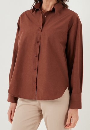 Kobieta ubrana w brązową koszulę na guziki z długim rękawem, zestawioną z beżowymi spodniami, stojąca na jednolitym jasnym tle.