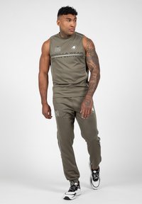 Olijfgroen mouwloos sporttop en bijpassende joggers met witte accenten en logo. De stof lijkt glad; de pasvorm is los en casual.