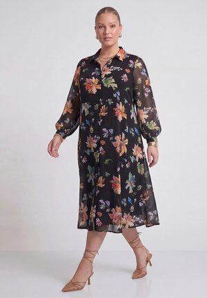 Kvinde, der modellerer en sort gennemsigtig midi-kjole med farverigt blomsterprint, lange pufærmer og tanfarvede snørelace spidse hæle mod en ensfarvet baggrund.