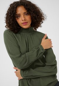 Blouse verte à manches longues avec un col haut smocké, un tissu texturé et des poignets boutonnés, coupe ample et détail plissé aux épaules.