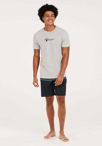 Grijze t-shirt met korte mouwen en zwart logo, gecombineerd met donkere zwemshorts met een gestreepte tailleband. Model staat blootsvoets.