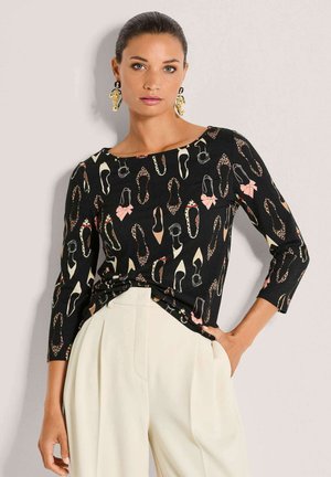 Schwarzes langärmeliges Oberteil mit einem Schuhdruck in verschiedenen Farben, einem Rundhalsausschnitt und einer verknoteten Detail am Saum. Kombiniert mit cremefarbenen Hosen.