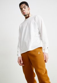 Weißes Langarmshirt mit Rundhalsausschnitt, kombiniert mit orange-braunen Jogginghosen, die auf dem linken Oberschenkel ein weißes Jordan-Logo aufweisen.
