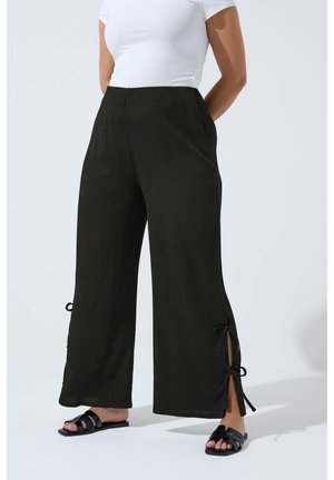 Donna che indossa pantaloni neri larghi con spacchi laterali e lacci, maglietta bianca a maniche corte e sandali neri aperti, in piedi contro uno sfondo semplice.