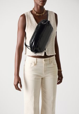 Borsa a tracolla in pelle nera con forma rettangolare e chiusura con cordoncino. Modello che indossa un gilet di maglia color crema e jeans chiari.