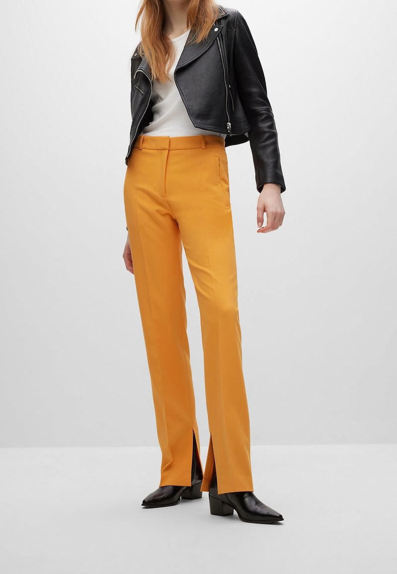 Femme portant un pantalon droit jaune moutarde avec des fentes à l'avant, des bottines noires, un haut blanc et une veste en cuir noire raccourcie.