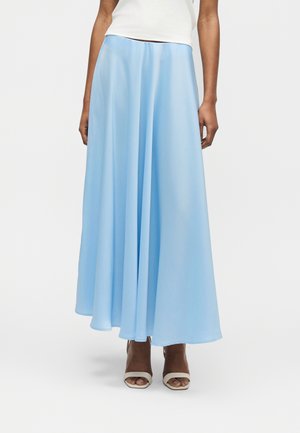 Γυναίκα που φοράει ροδαλή μακριά φούστα σε ανοιχτό γαλάζιο χρώμα, σε συνδυασμό με λευκά πέδιλα ανοιχτής μύτης και λευκό αμάνικο τοπ, στέκεται μπροστά σε απλό φόντο.