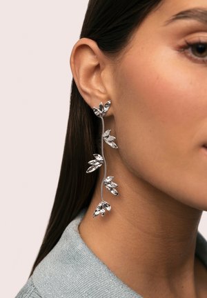 Pendientes en forma de hoja plateada con detalles brillantes, colgados de una cadena delicada. Sujetos a la oreja con un cierre de tuerca.