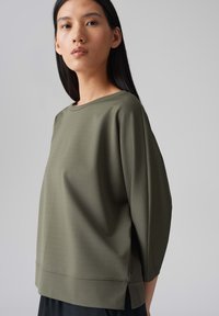 Olijfgroen, ruimvallend topje met een bootnek. Heeft driekwartmouwen en een afgeronde onderkant met zijsplitten voor extra comfort.